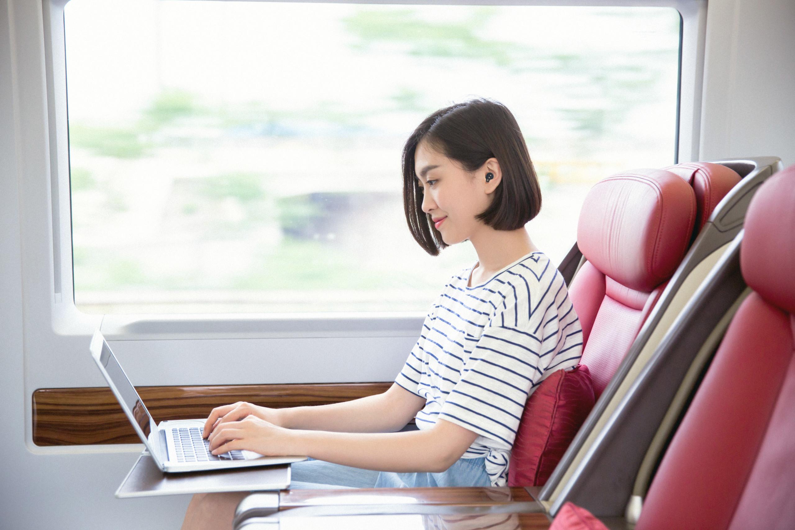 Woman sitting on train with laptop computer and earplugs.