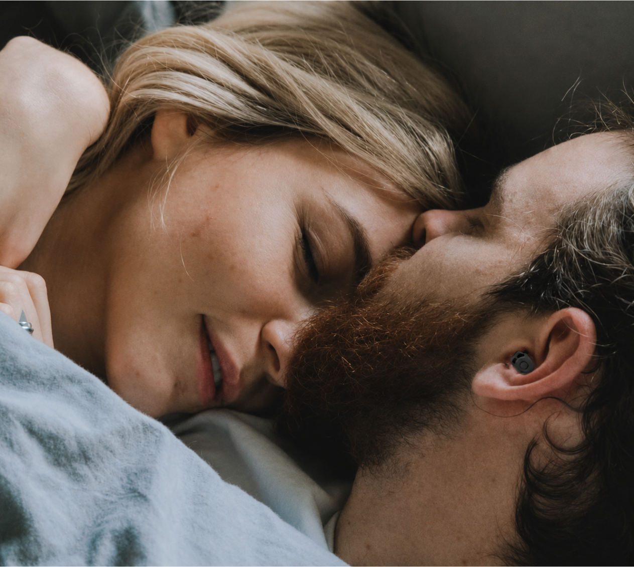 Couple sleeping peacefully together with man wearing QuietOn 4 sleep earbud — noise cancelling earbuds for undisturbed sleep beside a snoring partner